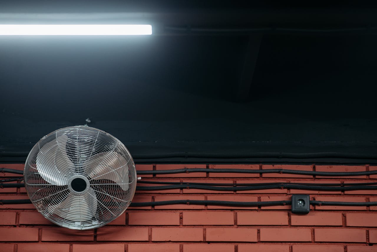 A close-up of a brick wall with a metal fan, wires, and fluorescent light. Industrial themed image.